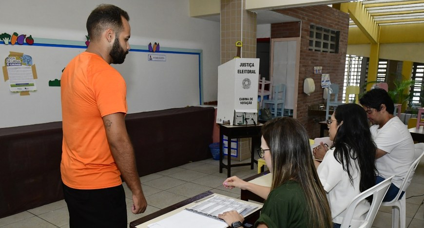 Registro fotográfico feito em uma seção de votação. O ambiente é uma sala de aula, com quadro br...