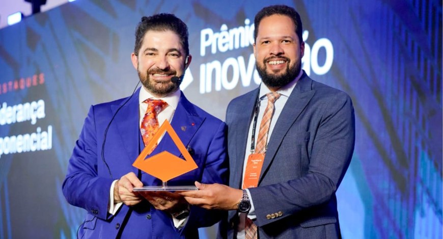 A imagem mostra dois homens usando ternos formais em um palco de premiação. Ambos seguram um tro...