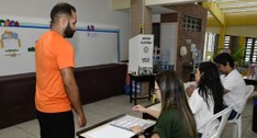 Registro fotográfico feito em uma seção de votação. O ambiente é uma sala de aula, com quadro br...