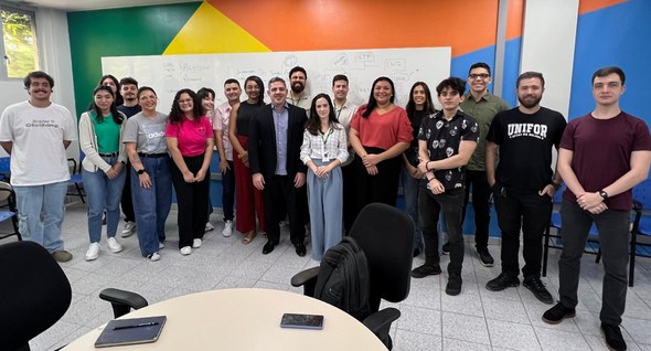 No registro, 19 pessoas posam em turma. Atrás, há uma lousa branca e uma parede com formas trian...