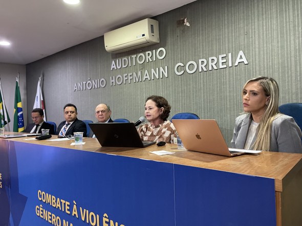 No registro, há uma bancada com um banner azul do evento. Atrás dela, há cinco pessoas sentadas ...