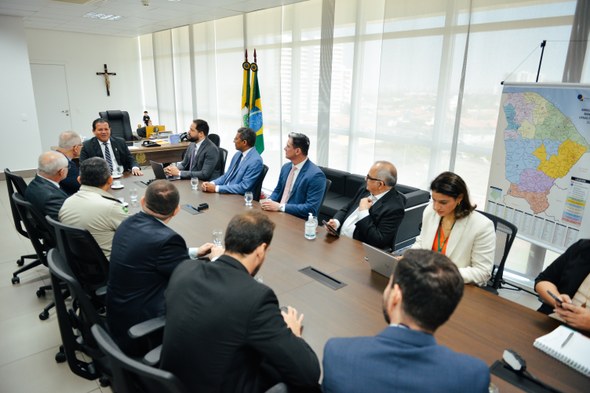 Na foto, ao redor da mesa de madeira, treze pessoas estão sentadas em cadeiras pretas, na sala d...