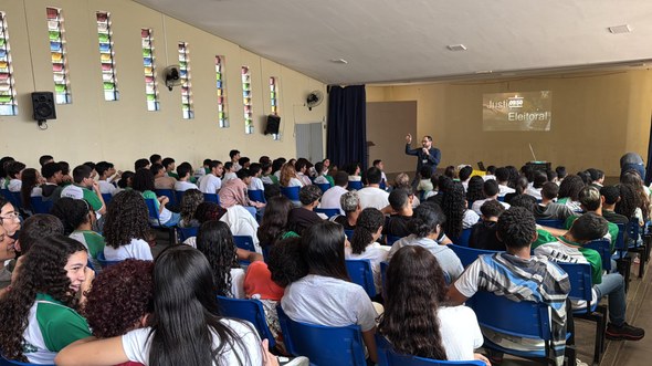 O registro foi feito em um auditório. Diversos alunos estão sentados em cadeiras azuis, olhando ...